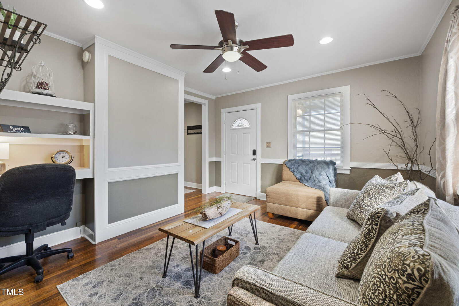 A cozy living room at 909 E F Street features a beige couch, patterned cushions, and a wooden coffee table. A ceiling fan spins above, while a desk with a chair sits in the corner. Nearby, a white door with a tall window overlooks the hardwood floor, partially covered by a rug.
