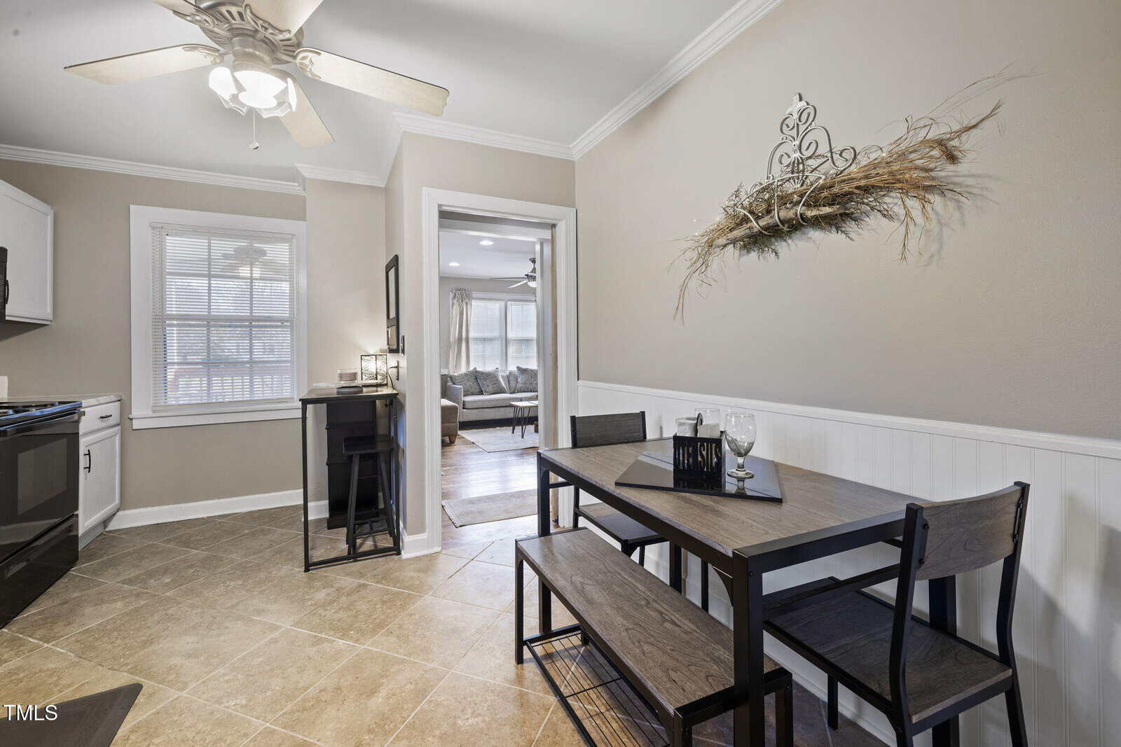 A kitchen area with a small dining table and two benches. There's a decorative wall piece above the table. The room has beige walls, a ceiling fan with lights, and tile flooring. A window and doorway lead to another room with a couch.