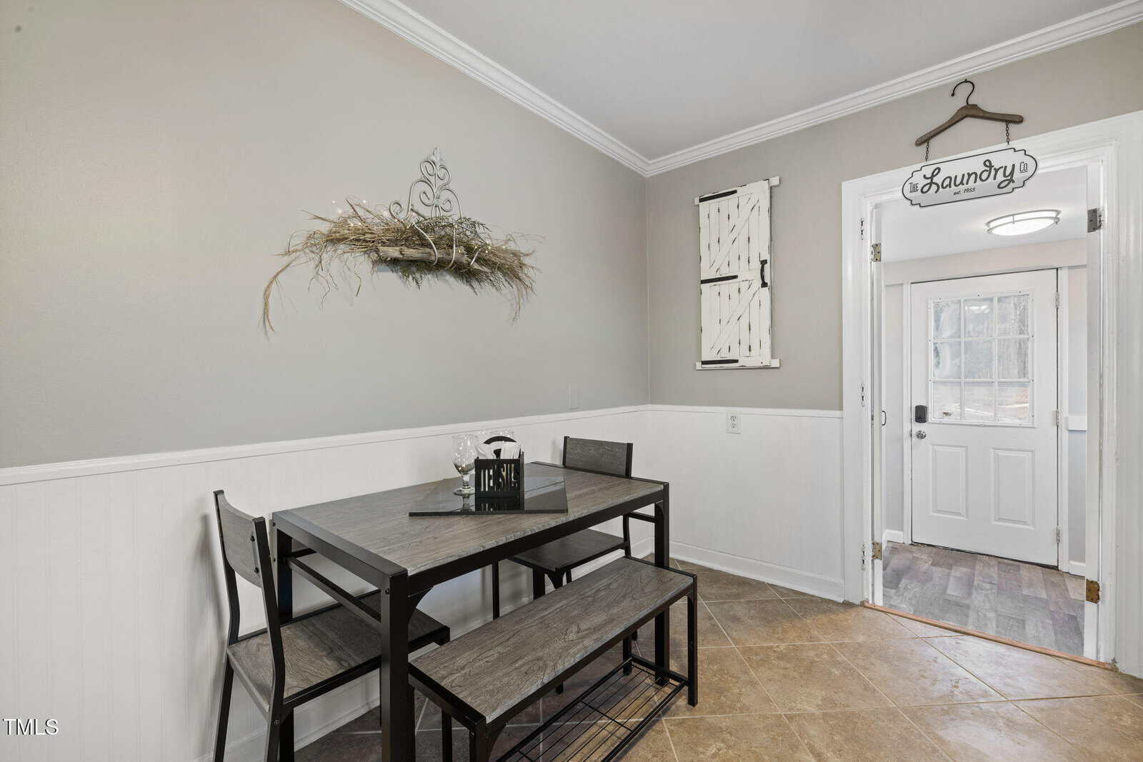 A small dining area with a wooden table and benches against a light gray wall. Above the table, there's a decorative branch arrangement. A door labeled "Laundry" leads to another room. The space has white wainscoting and beige tile flooring.