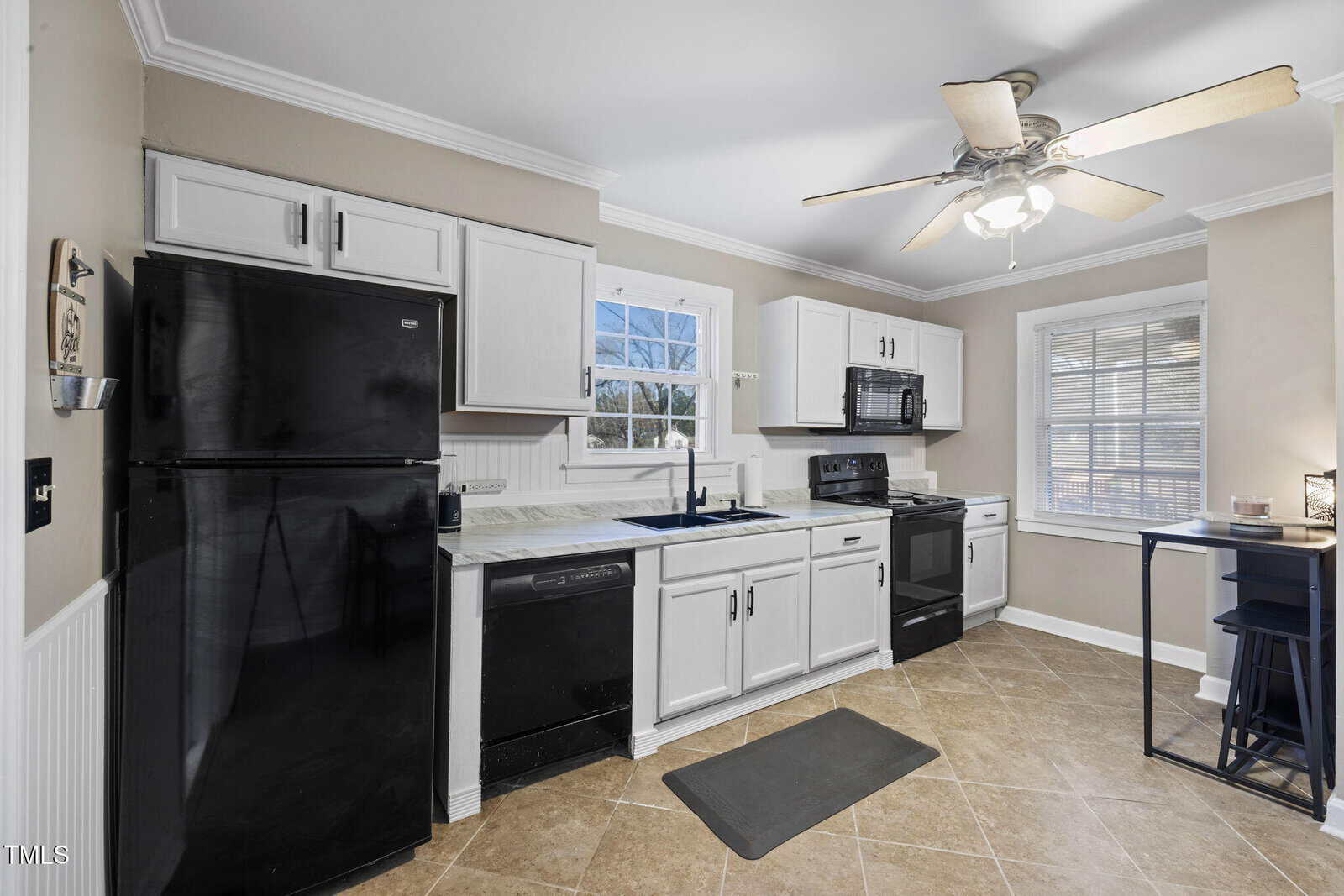 A modern kitchen features white cabinets, a black refrigerator, oven, and dishwasher. A ceiling fan hangs above, while a window provides natural light. The walls are light beige, and the floor is covered with beige tiles. A small table with stools is on the right.