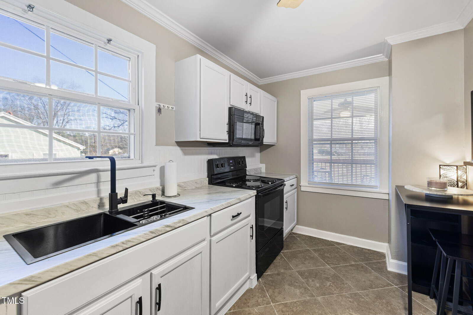 A small kitchen with white cabinets, black appliances, and a marble countertop. There is a window above the sink with a view outside, and another window to the side. A roll of paper towels is next to the sink, and bar stools are in the corner.