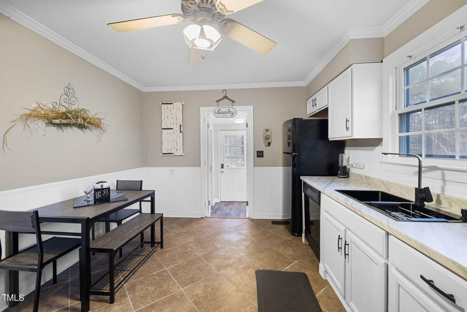 A kitchen with white cabinets, a black refrigerator, and a black sink. A small dining area features a black table with two benches. The walls are beige with white wainscoting, and there's a ceiling fan and wall decor. A door leads to another room.