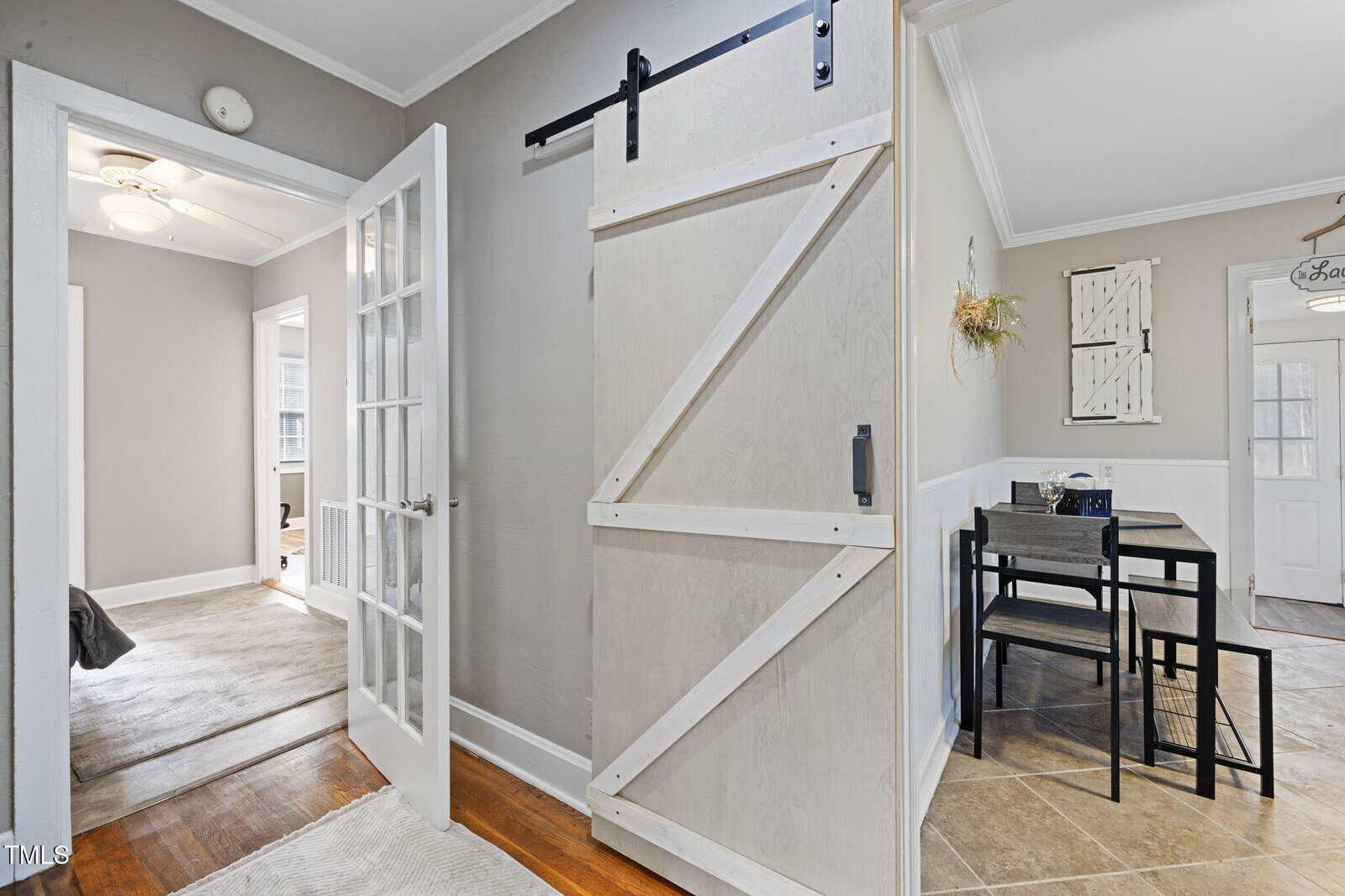 A bright hallway with light gray walls features a sliding barn door on the right. An open door on the left leads to another room. A small dining table and two chairs are visible in the adjacent room. The flooring is a mix of wood and tile.
