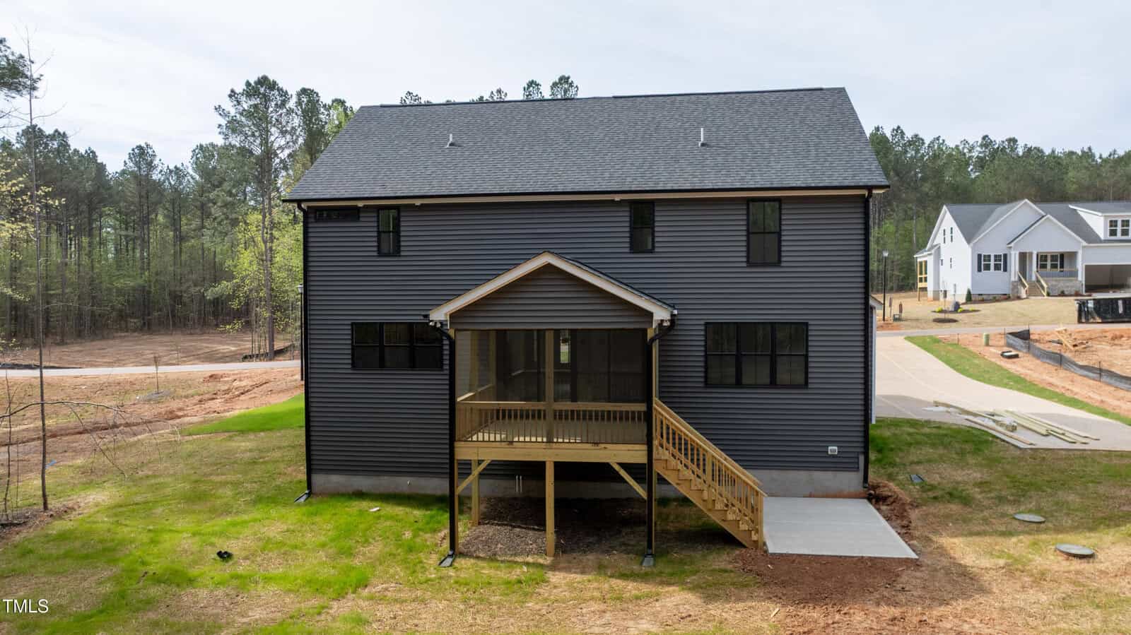 A two-story dark gray house with black windows and a covered back porch sits on a grassy lot. There is a small concrete walkway leading to the porch, and houses and trees are in the background.