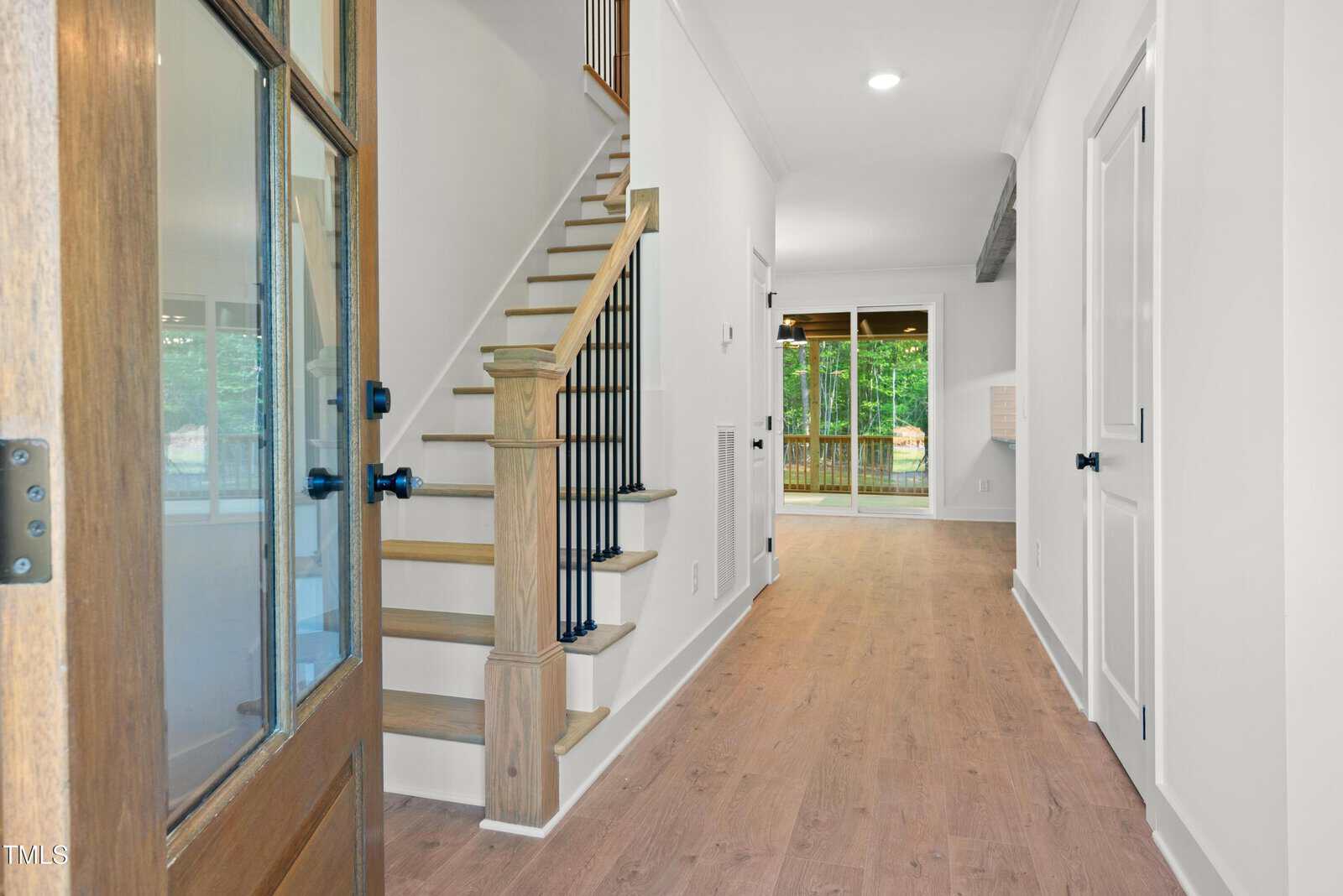 A view from the front door shows a hallway with light wood flooring, a staircase with wooden steps and black railings on the left, and a sliding glass door leading to a backyard at the end of the hall.