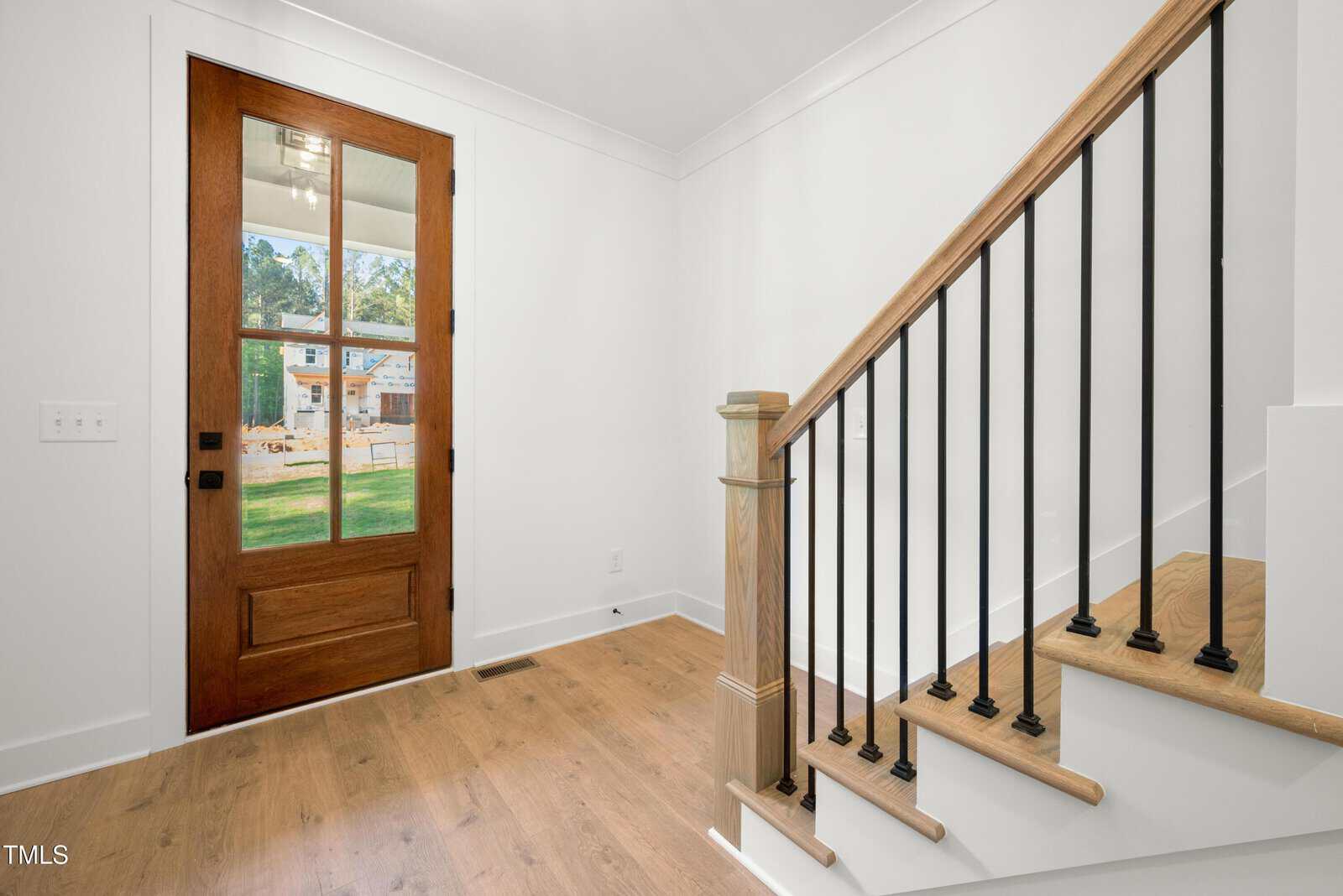 A wooden front door with glass panels and black hardware is beside a staircase with wooden steps and black metal railings in a bright entryway with light-colored wood floors and white walls.