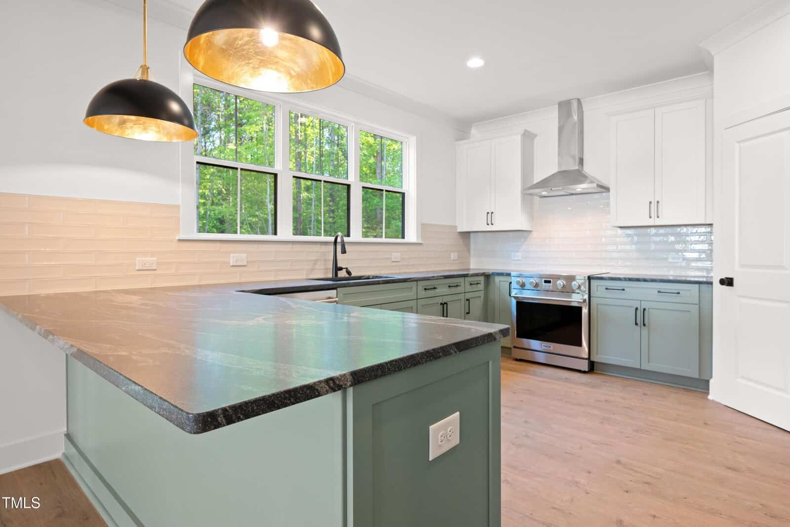 Modern kitchen with green cabinets, white upper cabinets, stainless steel appliances, black countertop, large windows, pendant lights, and light wood flooring. Subway tile backsplash in neutral beige tones.