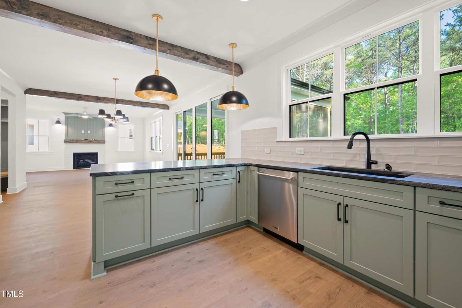 Modern kitchen with sage green cabinets, black countertops, stainless steel appliances, large windows, and pendant lights. Open layout leading to living area with fireplace and wood beams on the ceiling.