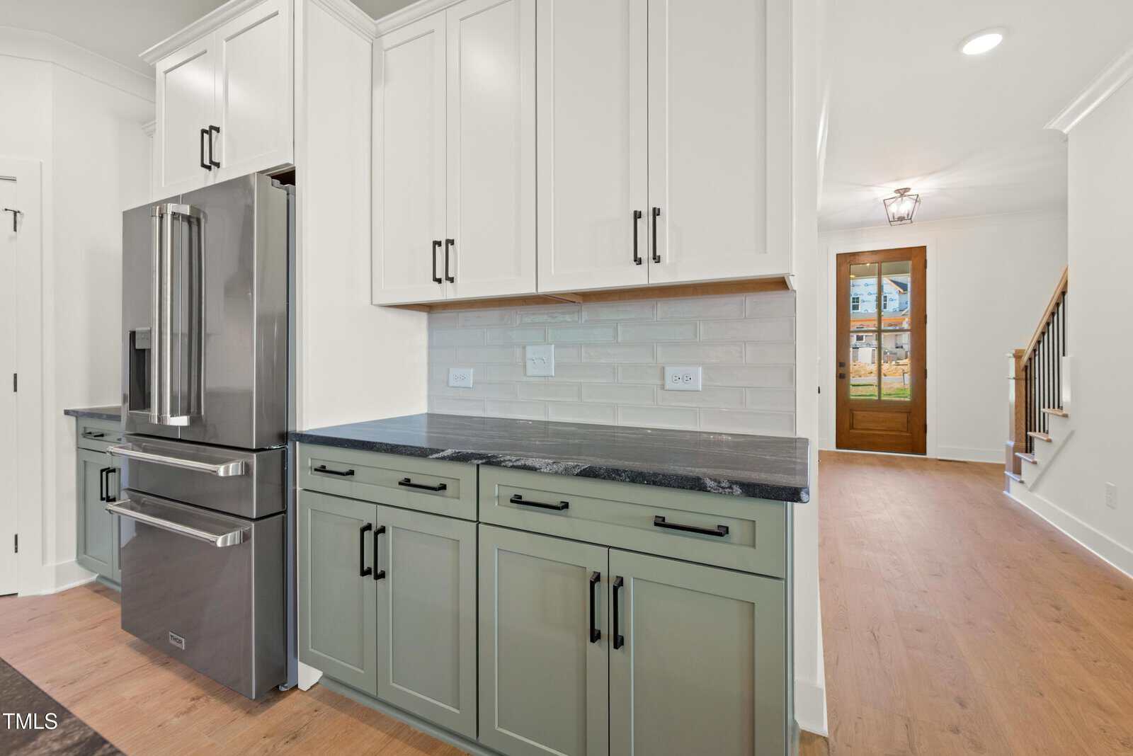 Modern kitchen with sage green lower cabinets, black countertops, white upper cabinets, stainless steel refrigerator, brick backsplash, and wood flooring. Hallway leads to a wooden front door and stairs.