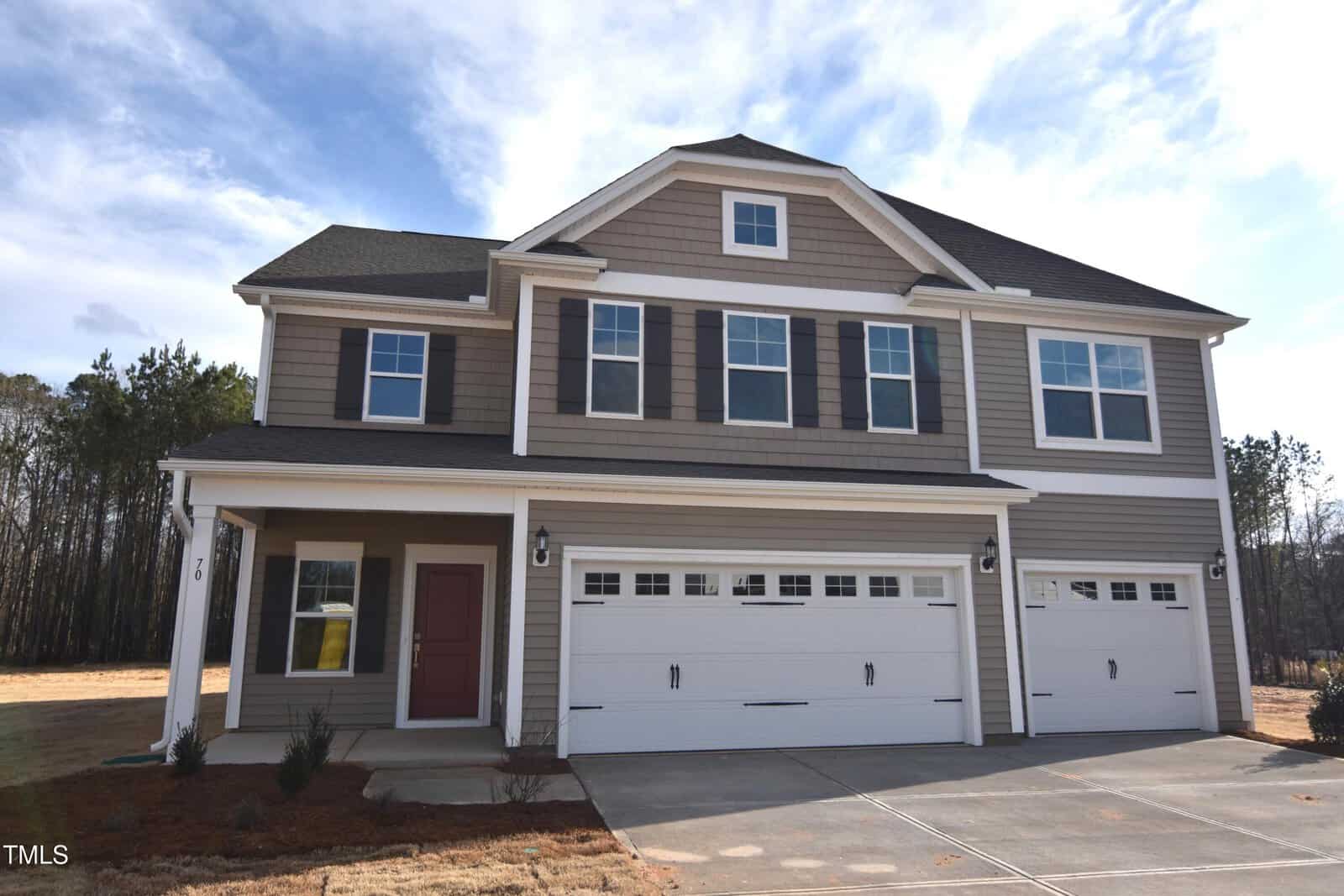 A two-story suburban house with beige siding, black shutters, and a covered front porch. The home features three white garage doors and a driveway, with a small lawn and trees in the background.