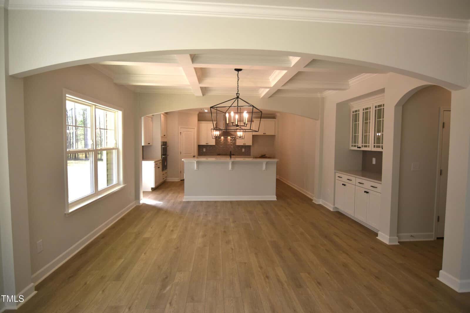Open-concept kitchen and dining area with light wood flooring, white cabinetry, modern pendant lighting, large windows, and coffered ceilings. Walls are light beige, and natural light fills the space.