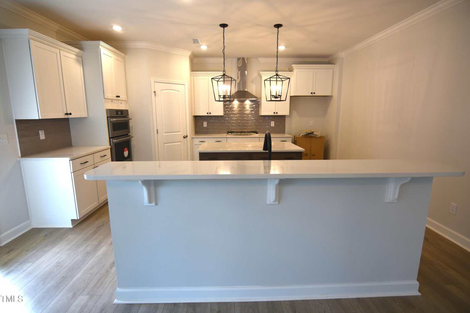 A modern kitchen with white cabinets, stainless steel appliances, two pendant lights over a white breakfast bar, wood flooring, and neutral-colored walls. The stove has a metallic backsplash and a hood above it.