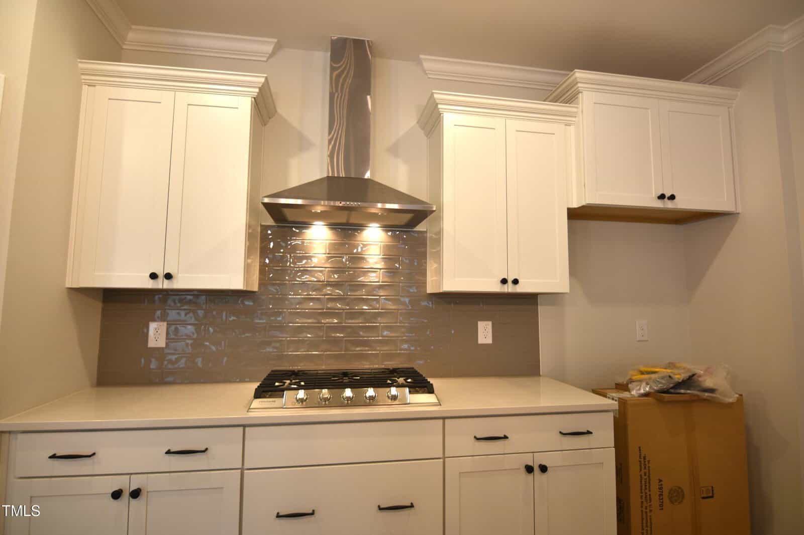 A modern kitchen features white cabinets, a stainless steel range hood with lights, a gas stovetop, a glossy gray tile backsplash, and a cardboard box with miscellaneous items on the right countertop.