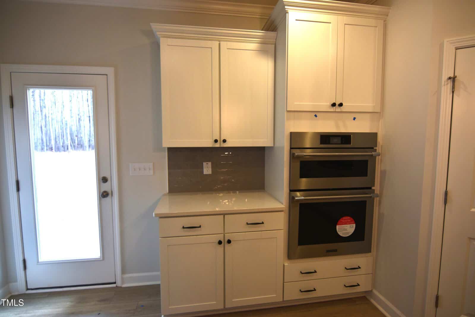 A kitchen area featuring white cabinets, a gray backsplash, a double wall oven, and a countertop. There is a glass door to the left, and light wood flooring runs throughout the space.