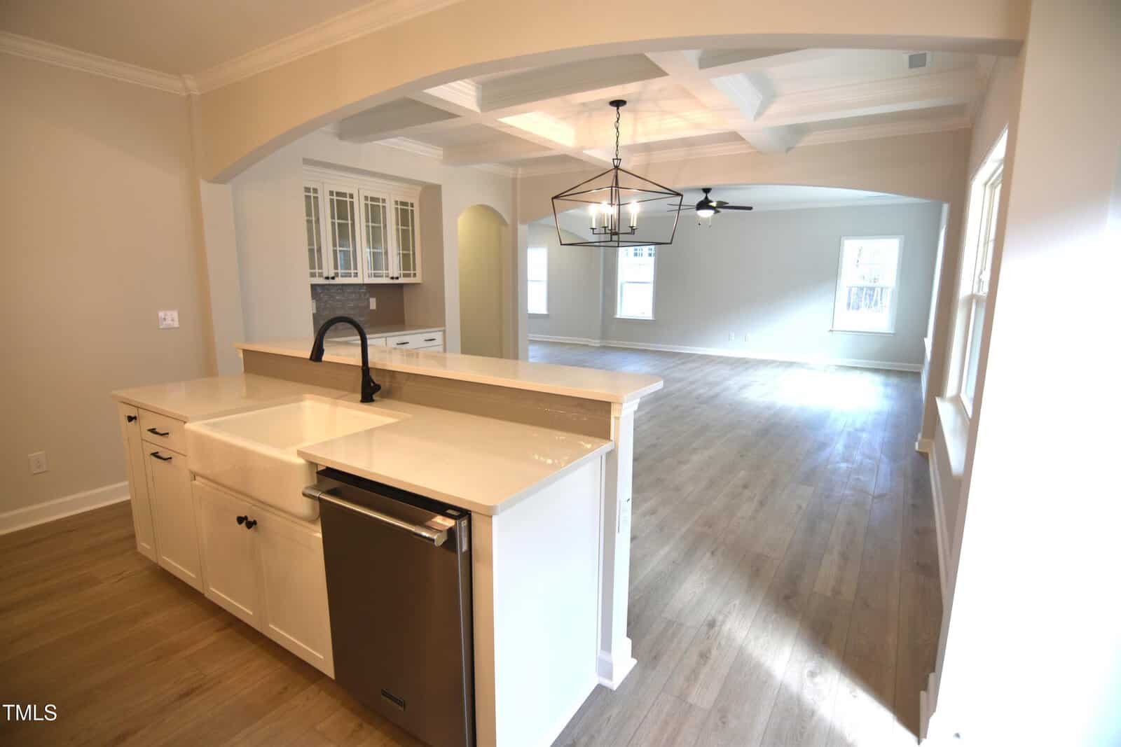 View from a kitchen with a farmhouse sink and dishwasher looking into an open living and dining area with wood floors, large windows, and a ceiling fan. White cabinetry and a light fixture are visible.