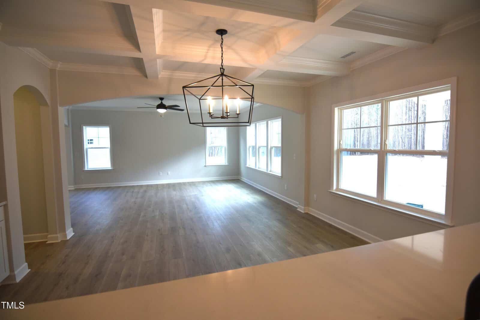 Spacious empty living area with light wood floors, large windows, a ceiling fan, and a modern black metal chandelier. White walls and a coffered ceiling create an open and bright atmosphere.