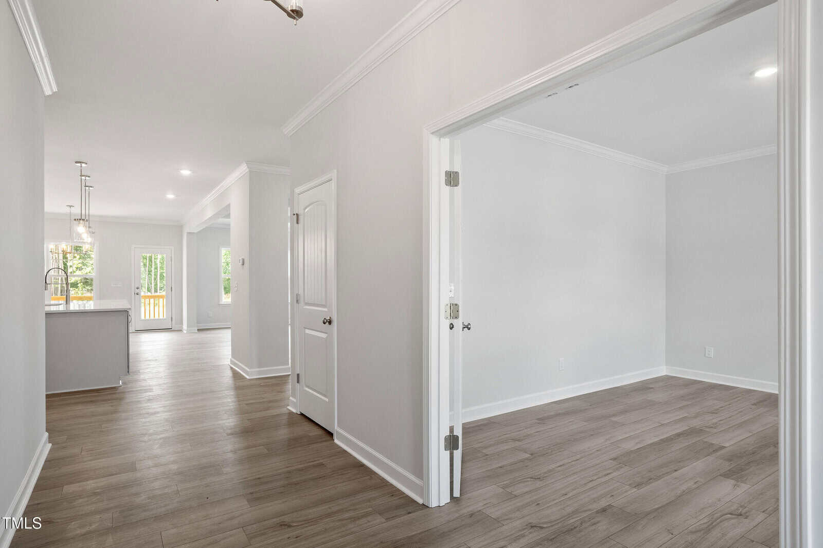 A view of a modern, empty home interior with light-colored walls and wood flooring. The image shows a hallway leading to an open kitchen, a closed door, and an open doorway to an empty room. Ample natural light enters from windows.