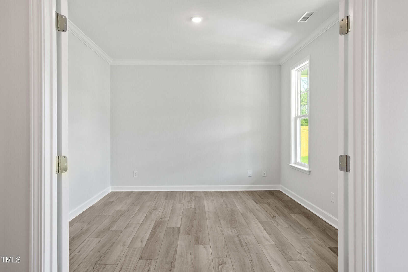 A bright, empty room with light-colored walls, wood-style flooring, white trim, a ceiling light, and a single window letting in natural light. Double doors with silver hinges are partially open in the foreground.