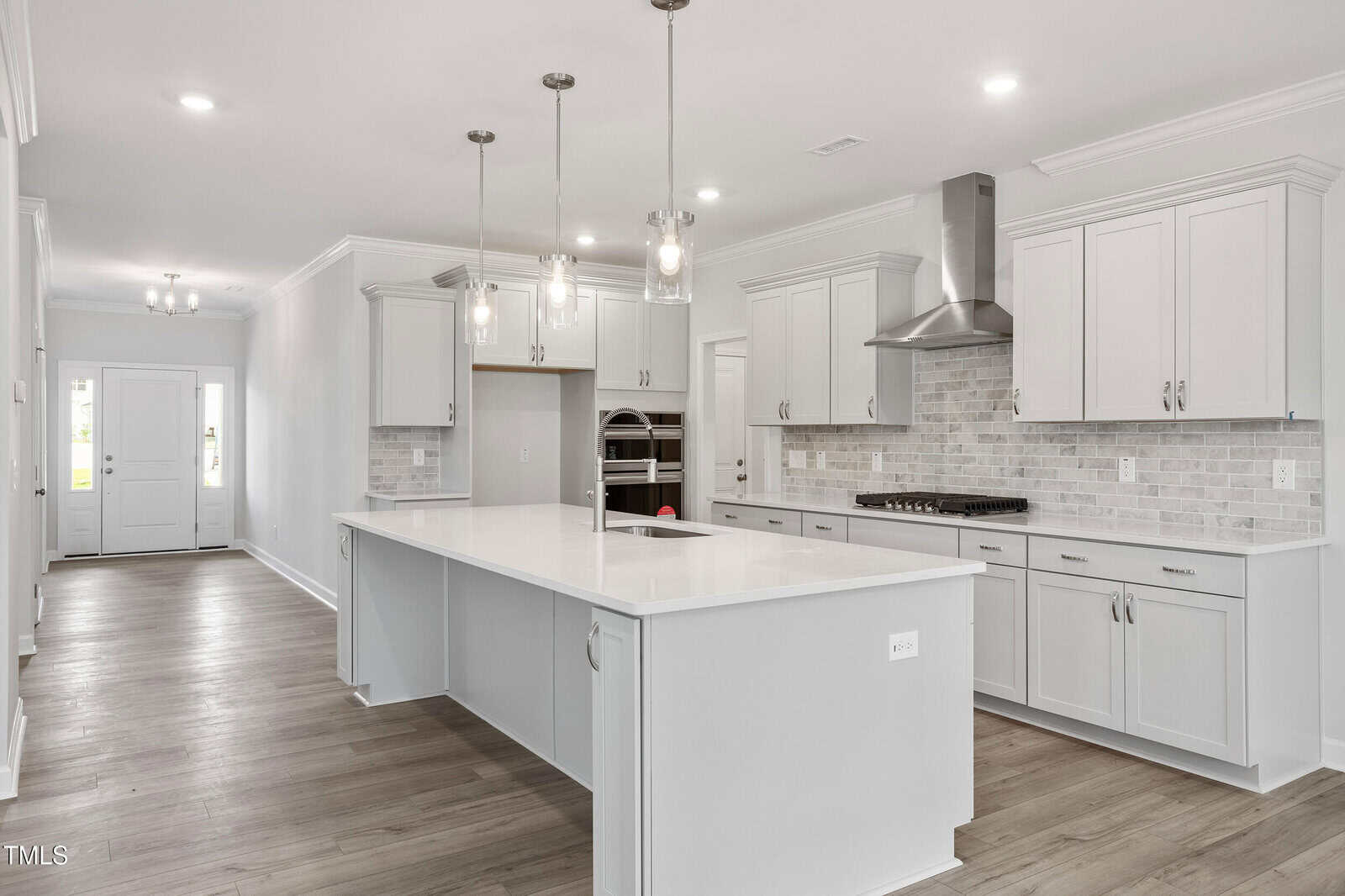 A modern kitchen with white cabinets, a large white island with sink, stainless steel appliances, pendant lights, light wood flooring, and a view of the front door down a hallway.