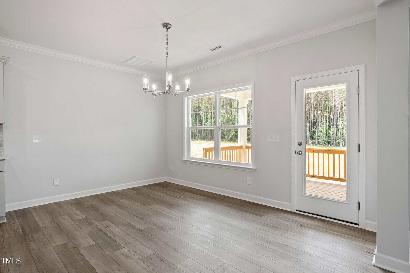 A bright, empty room with light wood-look flooring, a modern chandelier, large window, and a glass door leading to a deck outside with trees in the background. Walls are light-colored with white trim.