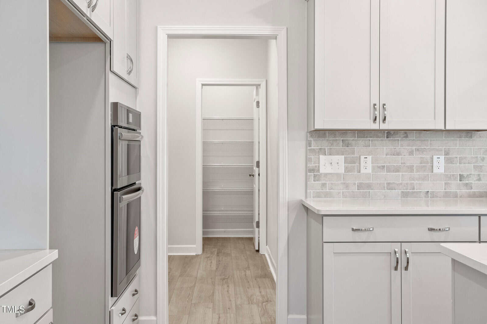 Modern kitchen with white cabinets, stainless steel double oven, light wood flooring, and a tiled backsplash. An open doorway leads to a walk-in pantry with multiple white shelves. Counters and wall outlets are visible.