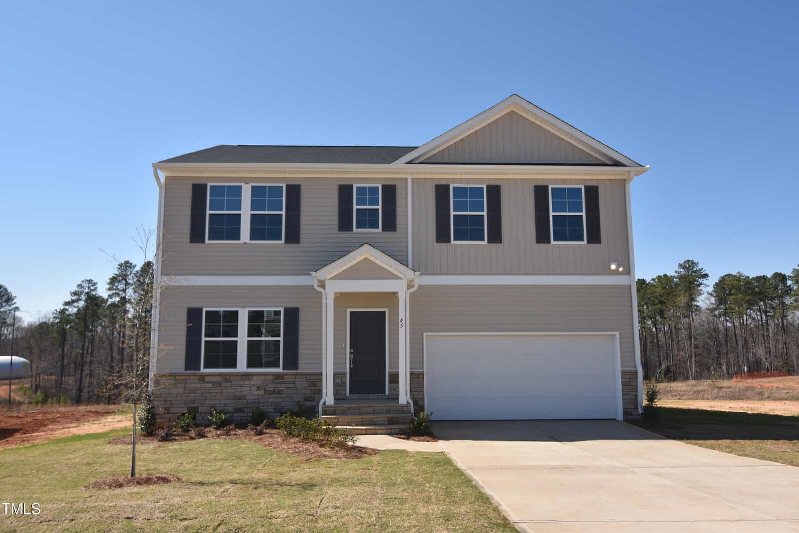 Two-story suburban house with beige siding, black shutters, a double garage, and a small front lawn. The home is set against a backdrop of trees and an unfinished yard.