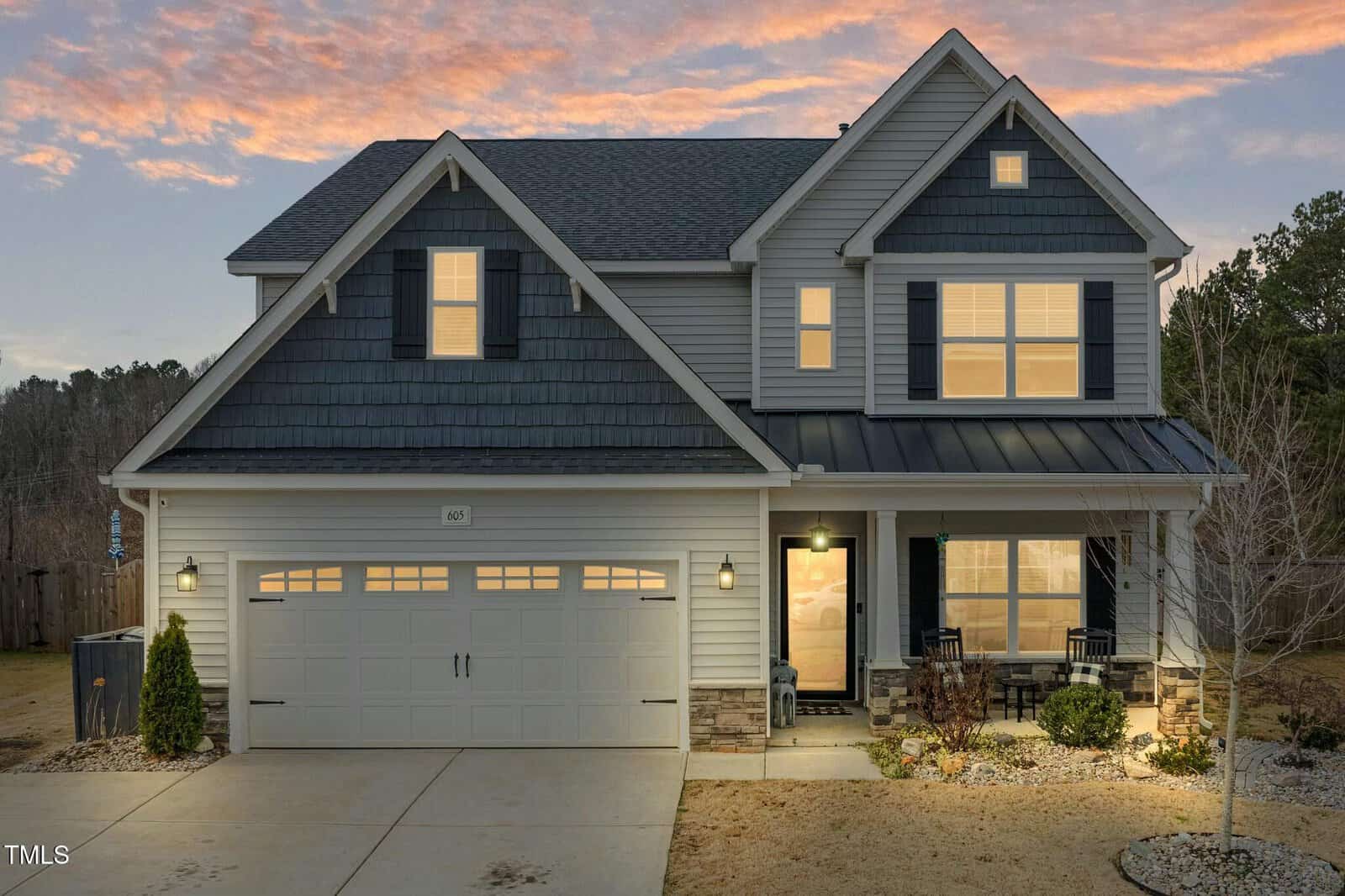 Two-story house with dark blue shingles and light gray siding, illuminated windows, a double garage, small front porch with two chairs, and a landscaped yard at sunset.
