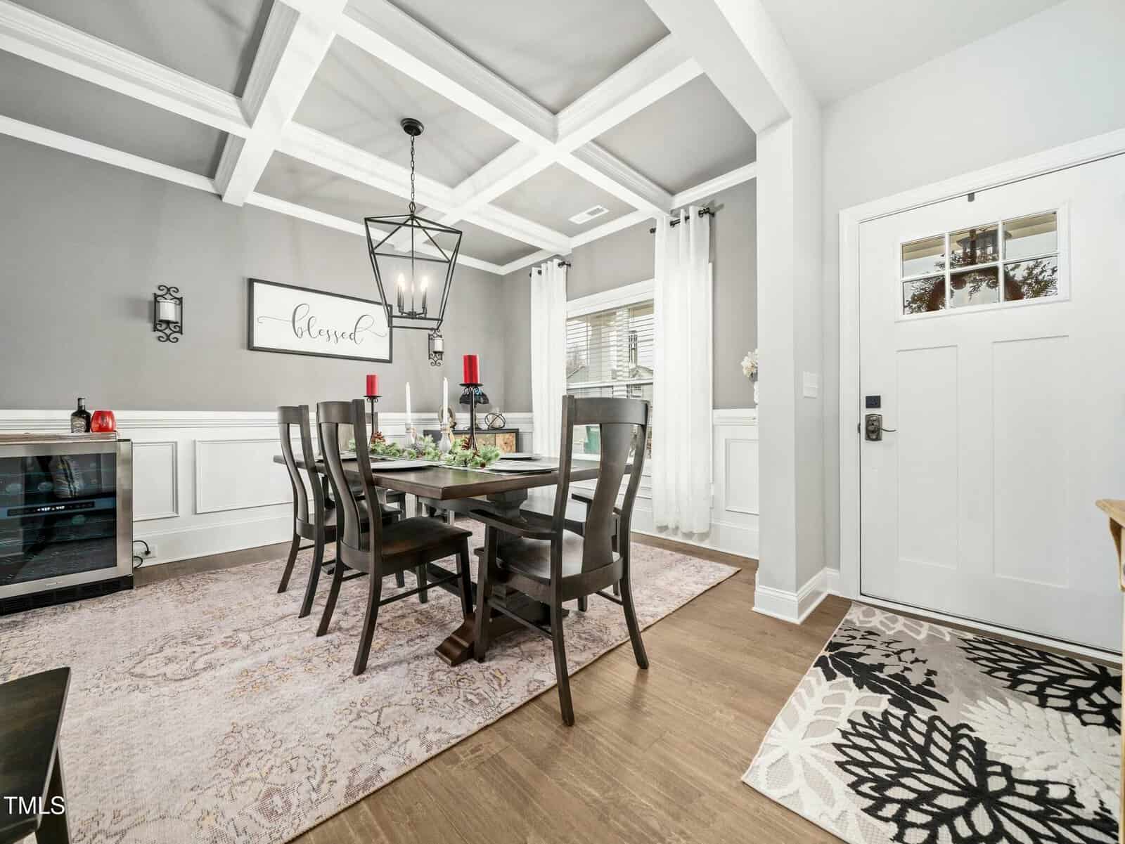 A modern dining room with gray walls, coffered ceiling, dark wood table and chairs, a patterned rug, and a sideboard. Sunlight enters from large windows; a door with glass panes is on the right, and a "Blessed" sign hangs on the wall.
