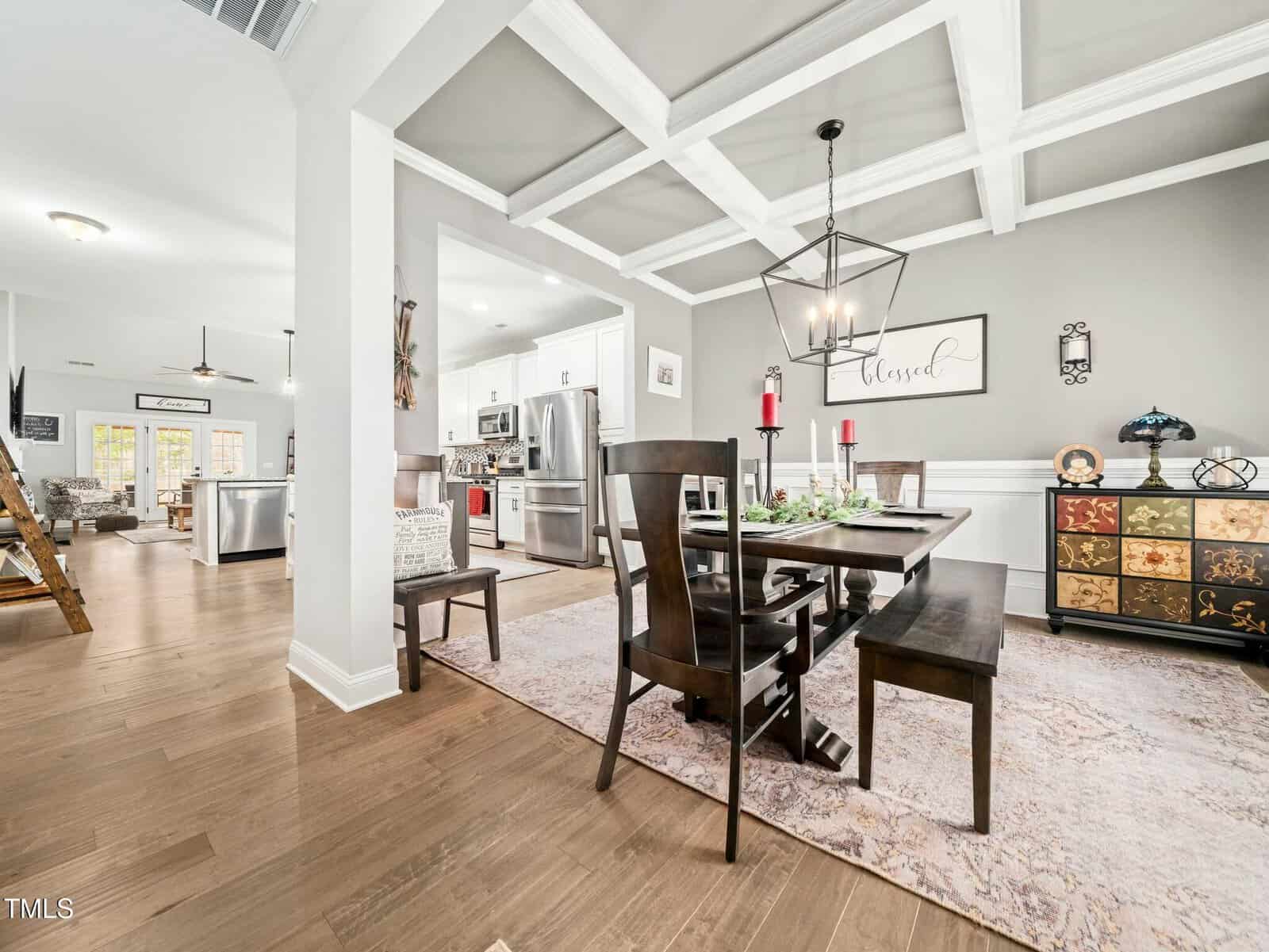 Modern dining room with dark wood table, chairs, and bench on a pastel rug. White walls, coffered ceiling, and a “blessed” sign. Kitchen with stainless appliances and island is visible in the background.