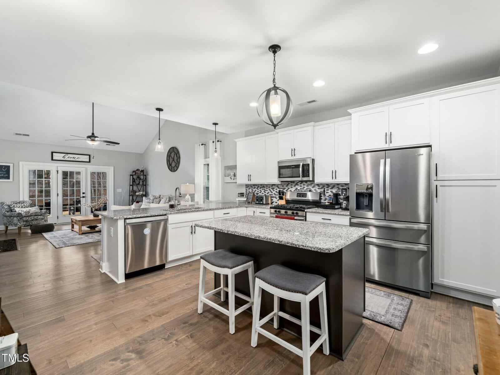 Modern kitchen with white cabinets, stainless steel appliances, an island with two stools, granite countertops, and hardwood floors. The open layout connects the kitchen to a living area in the background.