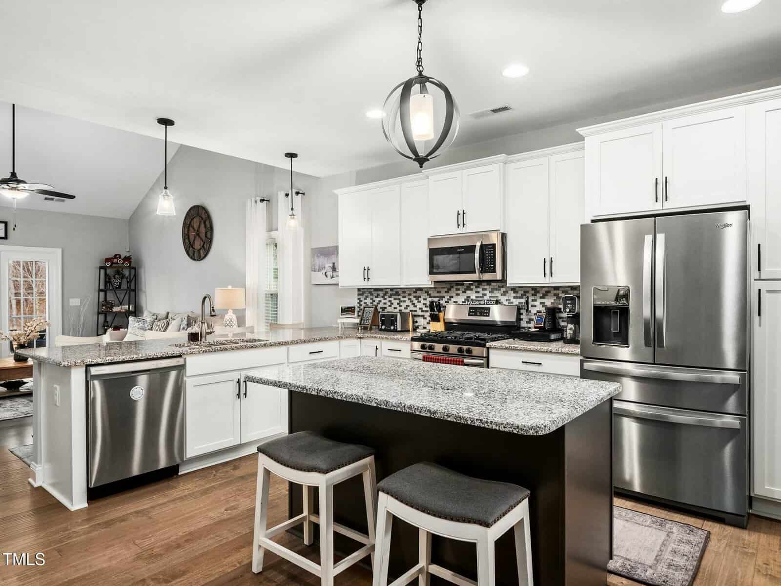 Modern kitchen with white cabinets, stainless steel appliances, a granite island with two stools, pendant lighting, and hardwood floors. An adjoining living room with couches and decor is visible in the background.
