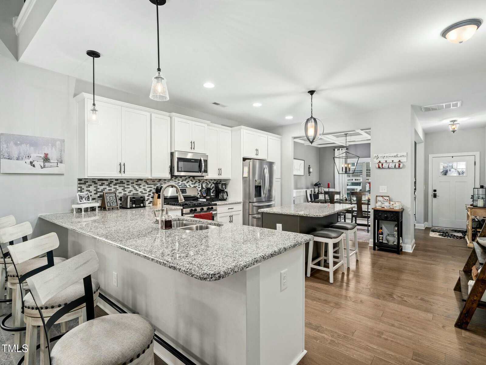 Modern kitchen with white cabinets, stainless steel appliances, granite countertops, bar stools, pendant lighting, and wood floors, opening into a dining area and entryway. Monochrome wall art and decor add cozy touches.