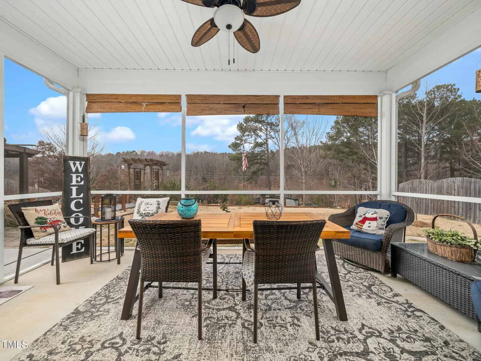 A screened-in patio with a ceiling fan, wicker chairs around a wooden table, benches with cushions, potted plants, a decorative "WELCOME" sign, and views of trees and a cloudy sky in the background.