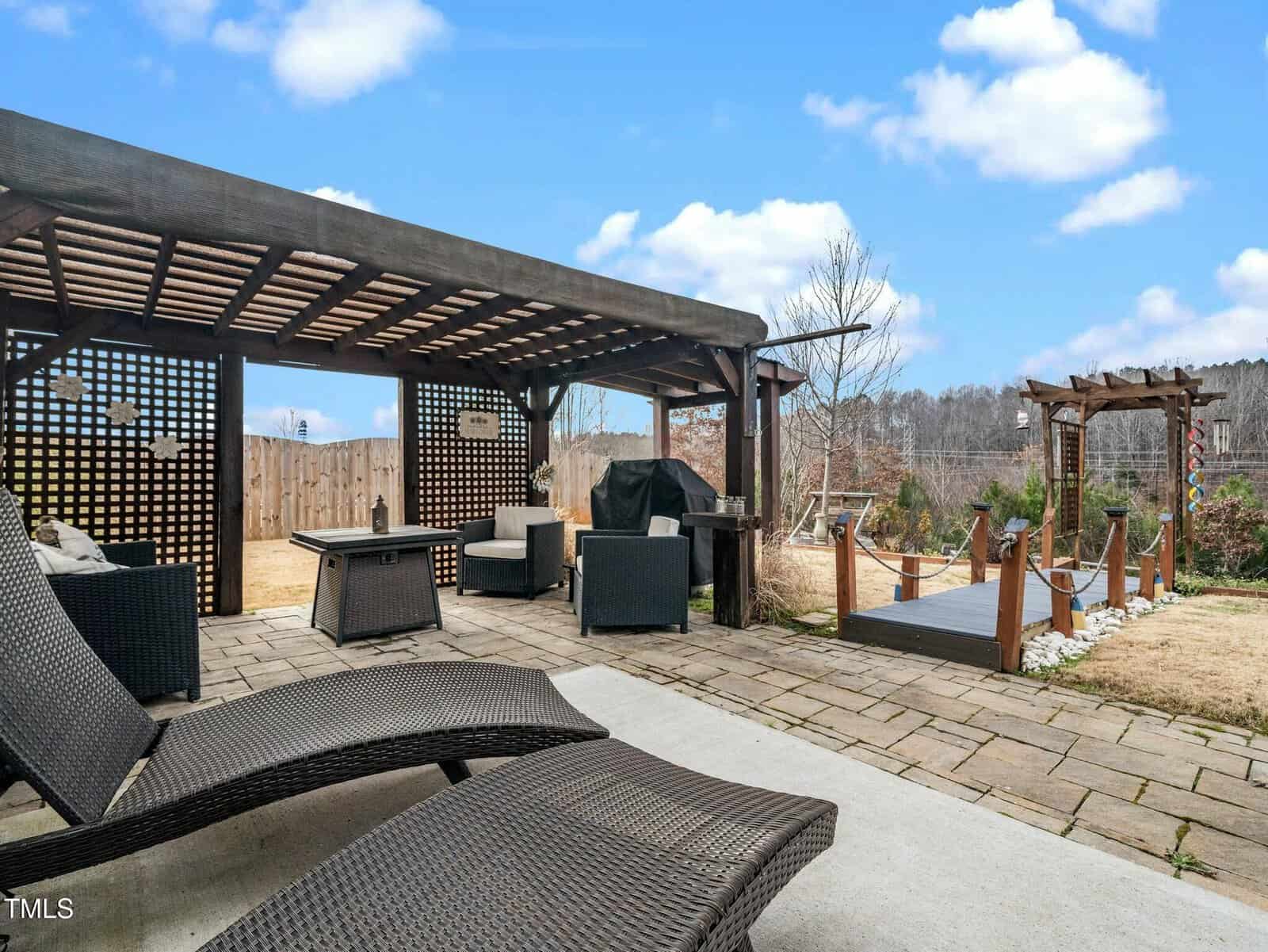 A backyard patio with wicker lounge chairs, a covered pergola, a grill, and a wooden bridge leading to a landscaped yard with a trellis, under a blue sky with scattered clouds.