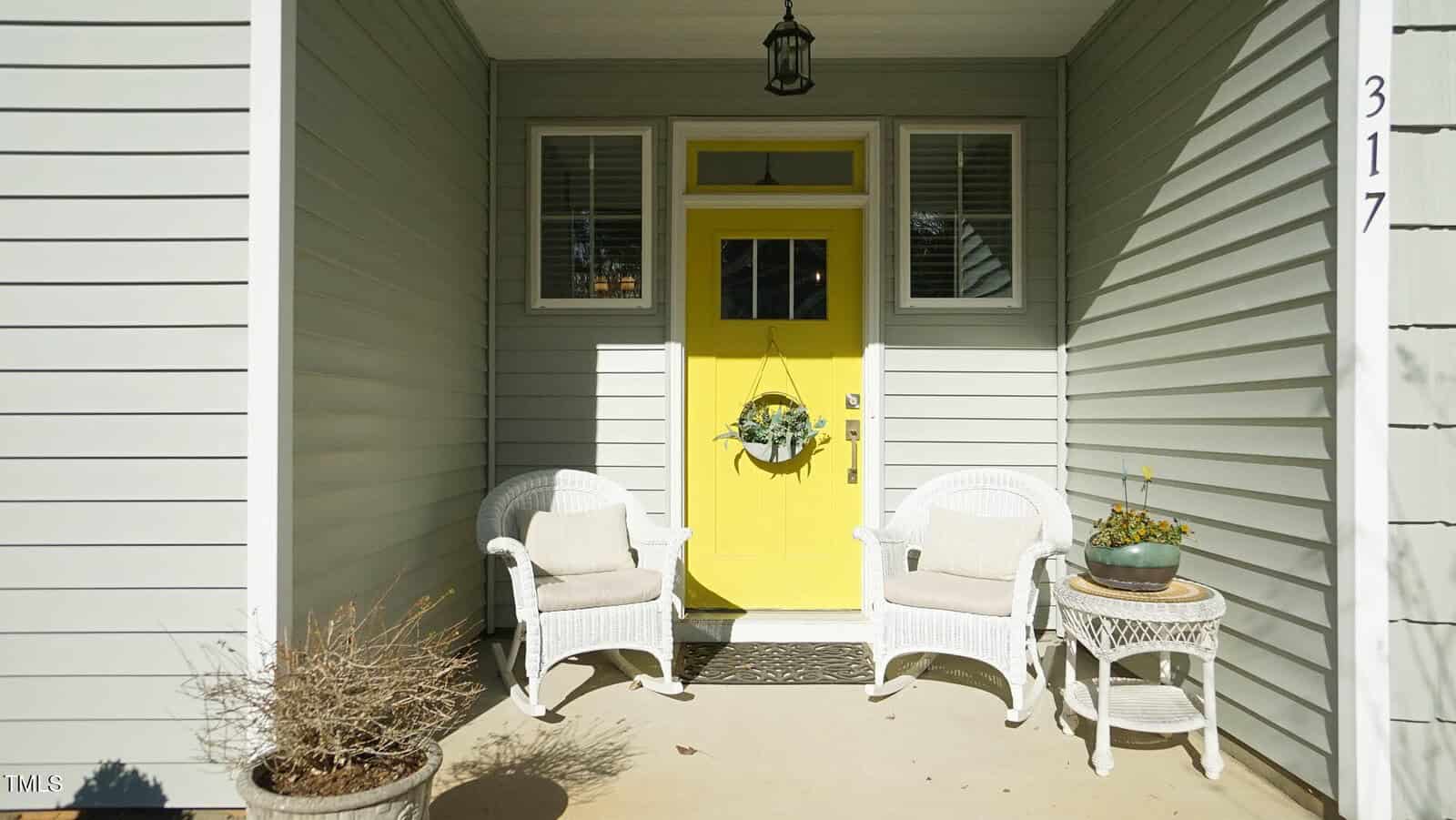 A covered porch with light gray siding features a bright yellow door, two white wicker chairs with cushions, a small white table holding a potted plant, and a dried plant in the foreground. House number 317 is visible.