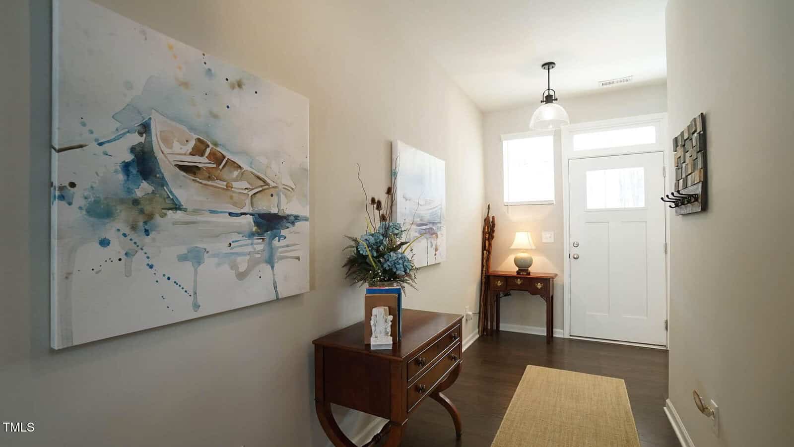 A hallway with light-colored walls, a painting of a boat, a wooden console table with a blue floral arrangement, a narrow rug, and a white front door with a window and transom letting in natural light.