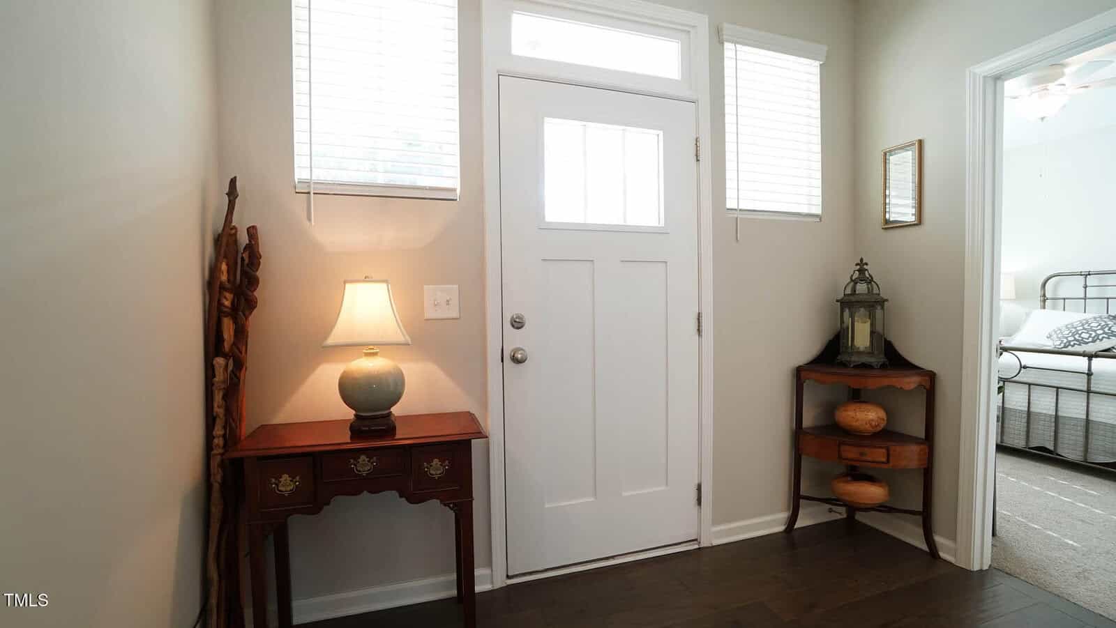A white front door with three square windows is flanked by two narrow windows. A small wooden table with a lamp is on the left, and a corner table with decorative items is on the right. A bedroom is visible through an open door.