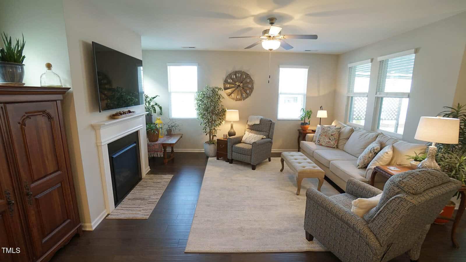 Bright living room with a sofa, two armchairs, a large wall-mounted TV above a fireplace, plants, lamps, and large windows letting in natural light. Neutral colors and wooden furnishings create a cozy atmosphere.