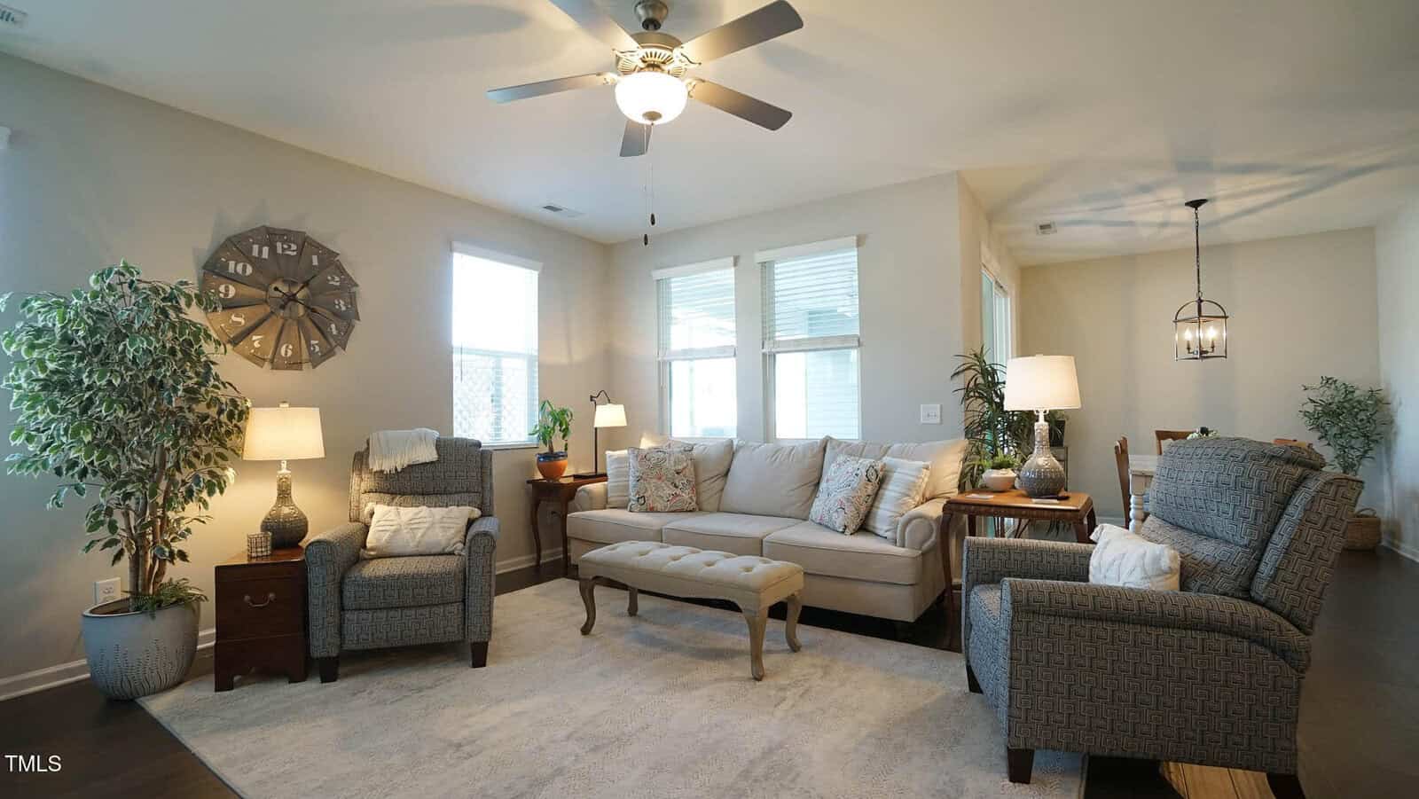 A cozy living room with a beige sofa, two armchairs, a bench, plants, lamps, and a large wall clock. Windows let in natural light, and a dining area is visible in the background.