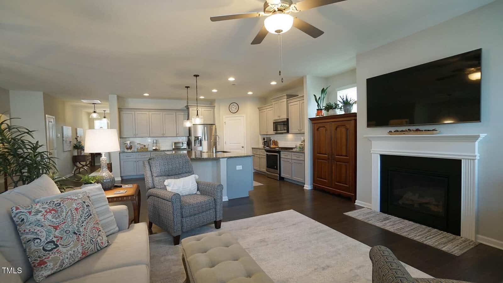 Open-concept living room and kitchen with gray cabinetry, a large TV above a fireplace, ceiling fan, armchairs, sofa, and a light rug on dark wood floors. Decorative plants and lamps add a cozy touch.