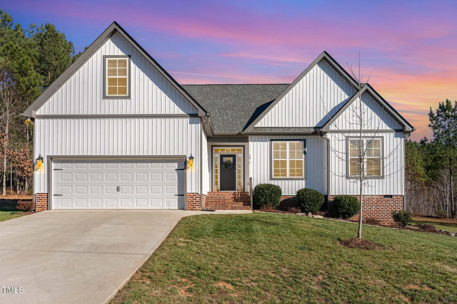 Single-story house with white vertical siding, brick accents, a two-car garage, front lawn, and a small tree. The sky shows a colorful sunset with purple, orange, and pink hues.