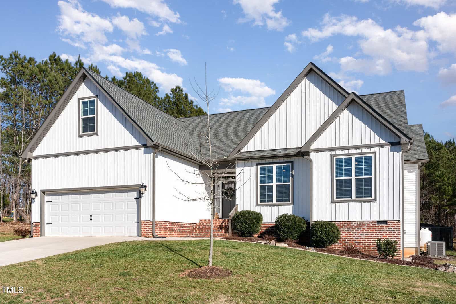 A modern single-story house with white siding and red brick trim, featuring a two-car garage, multiple windows, and a small tree in the front yard against a backdrop of trees and a blue sky.