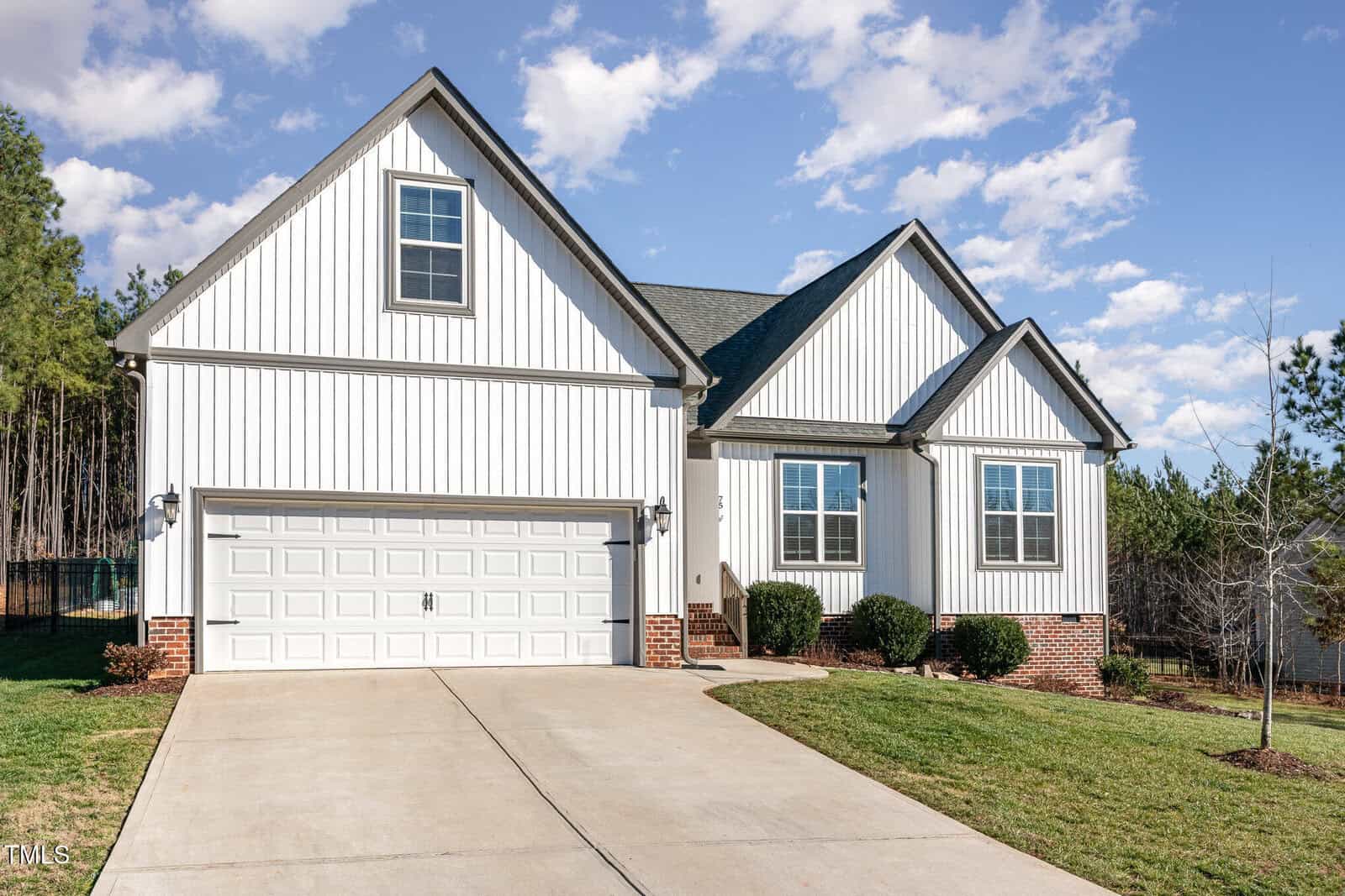 A modern, single-family house with white vertical siding, brick accents, and a two-car garage. The house has a sloped roof and a concrete driveway, with grass and small shrubs in the front yard.