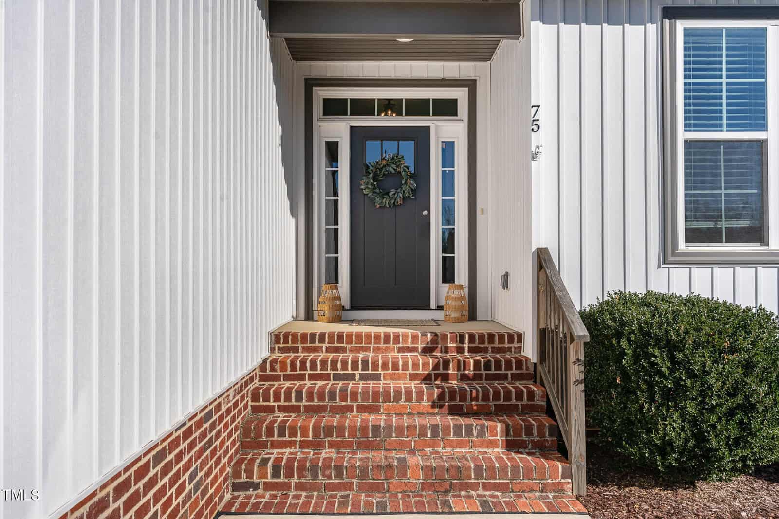 A brick staircase leads to a dark gray front door with a wreath. There are two decorative items on either side of the door. The house exterior features white vertical siding and a window with shutters to the right.