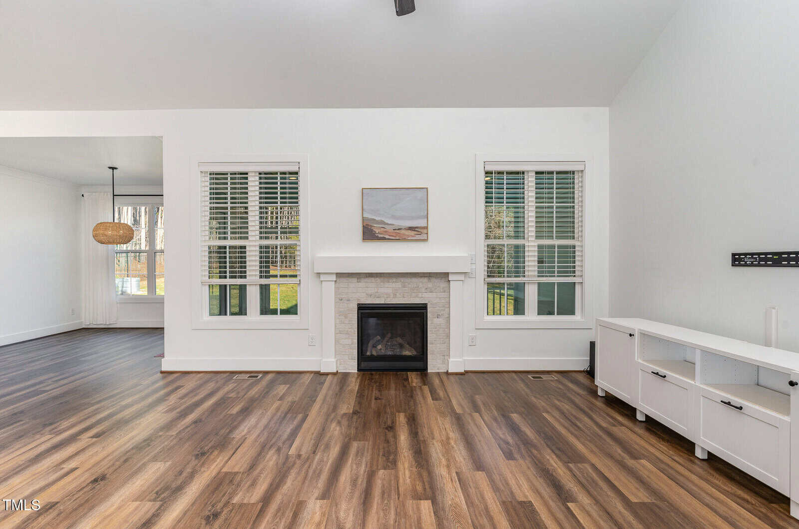 Bright living room with wood floors, white walls, two large windows, a fireplace with a painting above it, and a white media console. Dining area with hanging light is visible to the left.