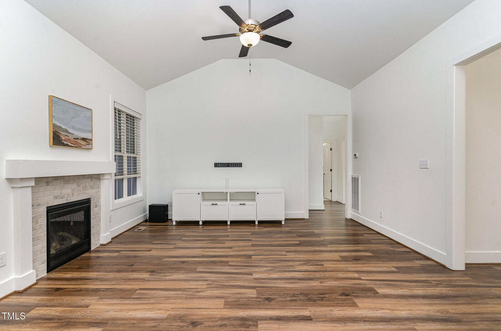 Modern living room with vaulted ceiling, ceiling fan, wood flooring, white walls, fireplace on the left, TV stand against the far wall, and two doorways on the right. Large window by the fireplace.