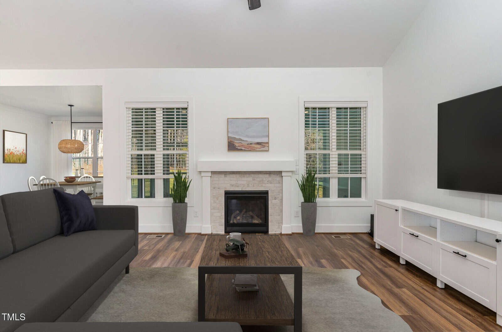 Modern living room with a black sofa, coffee table, TV, white media console, fireplace, plants, and wood flooring. Dining area visible in the background. Walls are white with two windows and a painting above the fireplace.