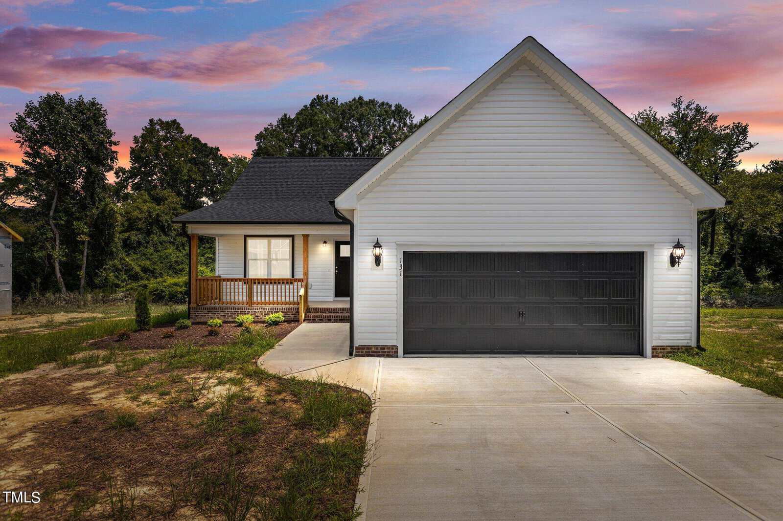 A modern single-story house with white siding, a black garage door, and front porch sits on a concrete driveway. The yard is partially landscaped, and trees are visible in the background at sunset.