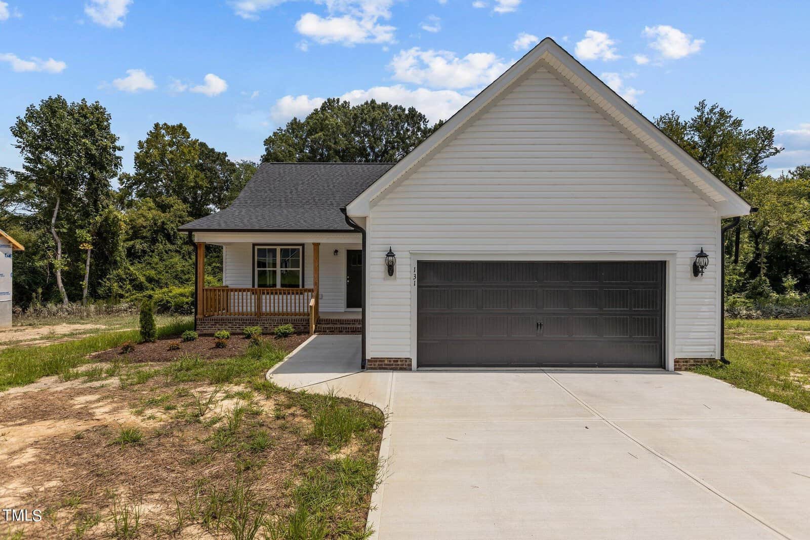 A single-story house with white siding, a dark gray two-car garage door, a covered front porch with wooden railings, and a concrete driveway. The yard has minimal grass and some small landscaped bushes.