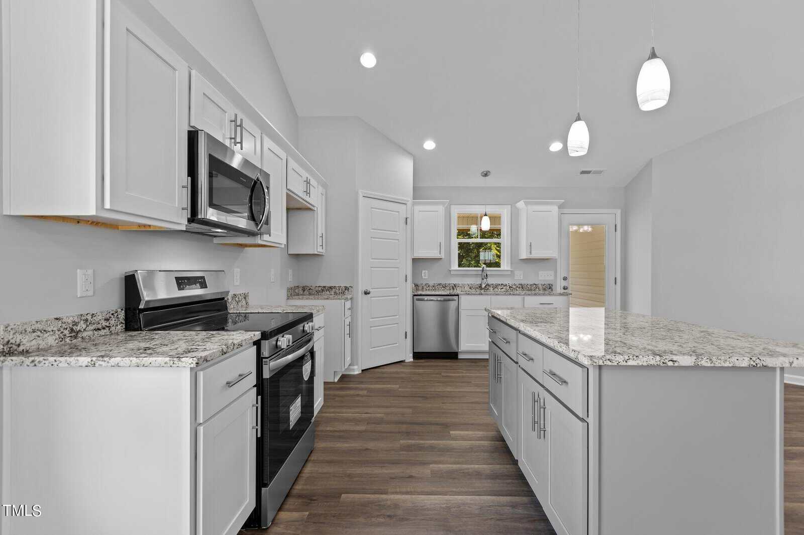 A modern kitchen with white cabinets, stainless steel appliances, granite countertops, a large island with drawers, pendant lights, and wood flooring. There is a window above the sink and a white door in the background.